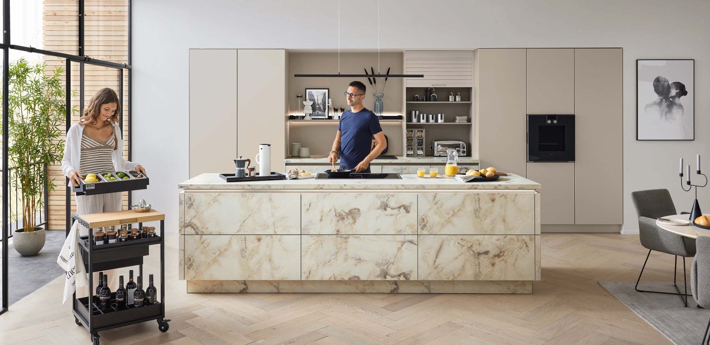 Großzügige Ballerina Küche mit zentraler Kochinsel in Marmoroptik, Mann beim Kochen und Frau mit Servierwagen, modernes Wohnkonzept mit offenem Essbereich bei Bodensee Küchen in Markdorf