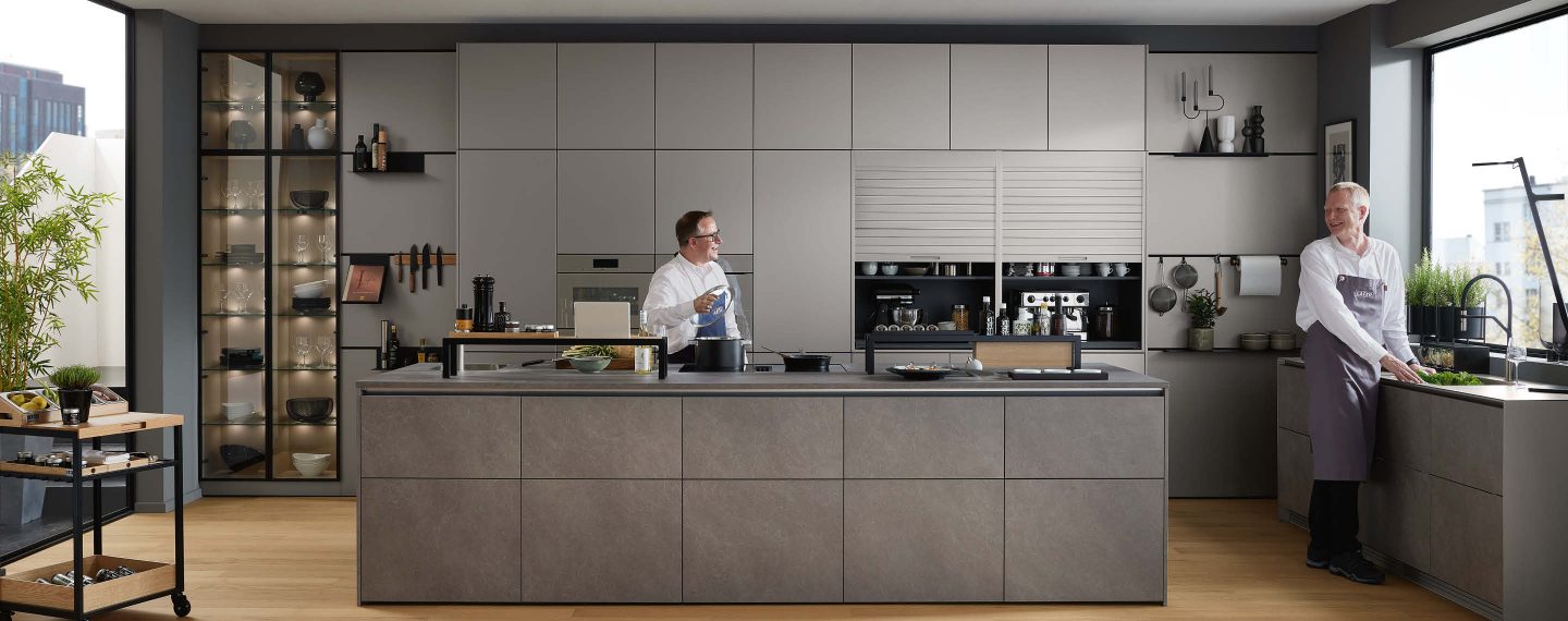 Moderne Designküche in gedeckten Grautönen mit großzügiger Kochinsel, integrierter Vitrine und offenen Funktionsbereichen – ideal für gemeinsames Kochen und professionelle Küchenabläufe