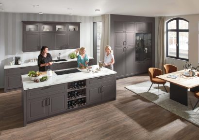 Klassische Landhausküche in dunklen Tönen mit Kochinsel und integrierter Weinkühlung, drei Frauen bereiten gemeinsam Speisen zu in gemütlicher Wohnküche mit Essbereich