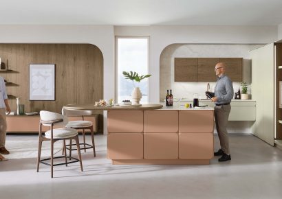 Moderne Ballerina Küche mit Kochinsel in warmen Natur- und Rosétönen, Mann bei der Zubereitung von Getränken, kombiniert mit integriertem Essbereich und stilvollen Holz- und Designakzenten