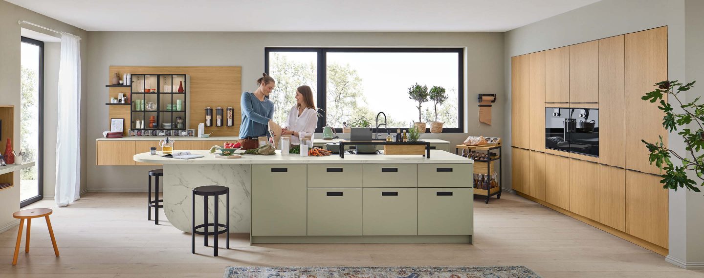 Einbauküche im Küchenstudio mit Kücheninsel, Holzfronten, Hochschränken und integrierten Geräten, zwei Personen bereiten Speisen an der Arbeitsfläche zu